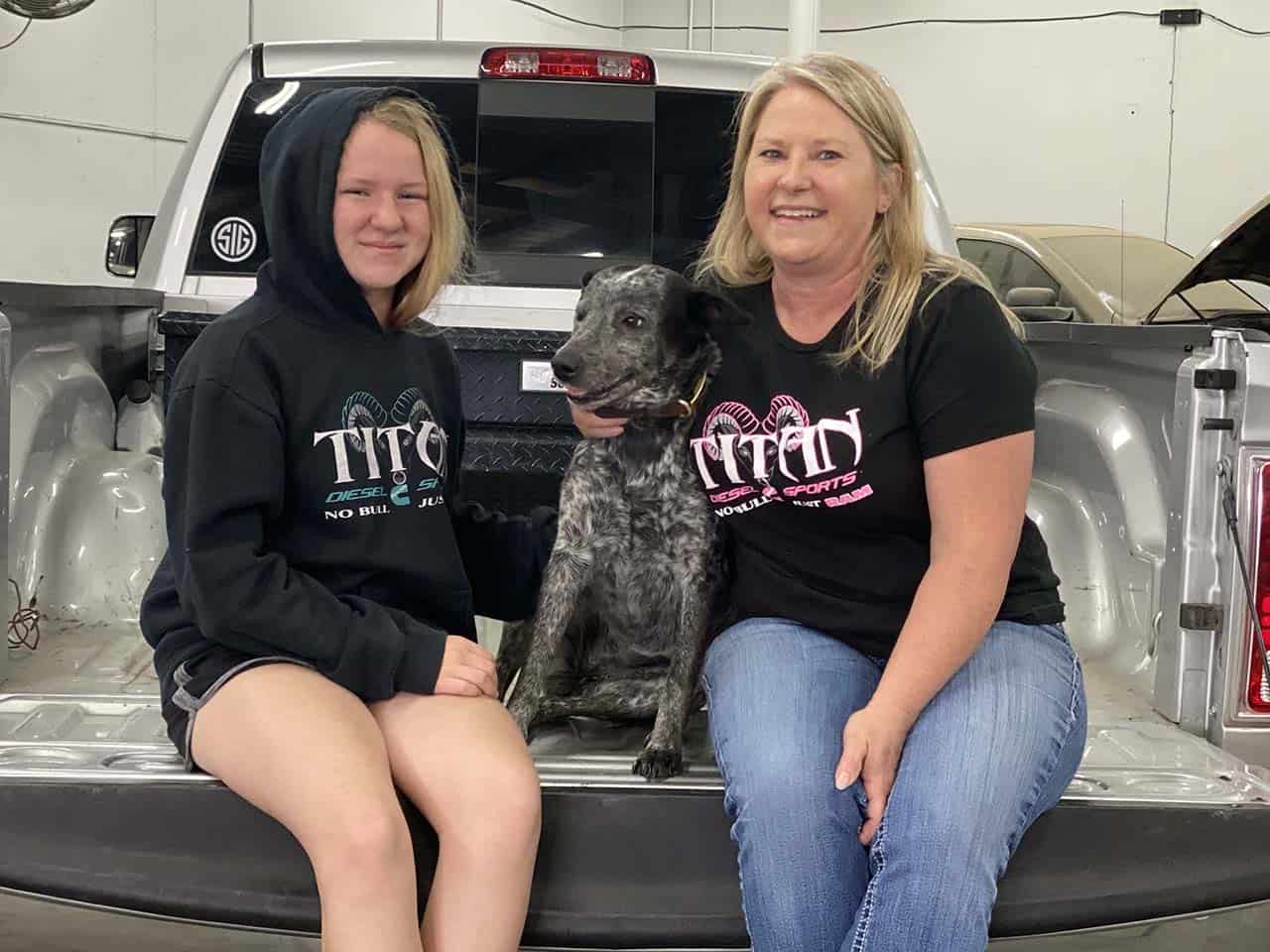 Woman and girl sitting on the bed of a pickup truck with a dog.